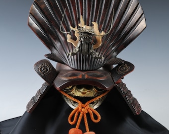 May include: A traditional Japanese samurai helmet, or kabuto, with a black and red lacquered finish. The helmet features a decorative dragon crest and a black and gold face mask. The helmet is displayed on a black cloth with orange tassels.