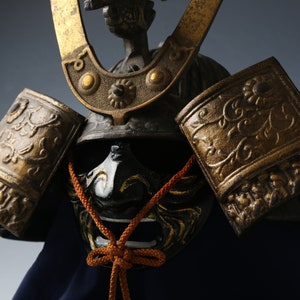 Op de afbeelding: Een close-up van een traditionele Japanse samurai-helm met gouden accenten en een zwart gezichtsmasker. De helm is versierd met ingewikkelde bloemmotieven en heeft een rood en wit touw dat om het masker is gebonden.