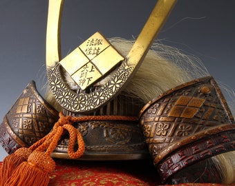 May include: A miniature samurai helmet with intricate details, including a gold-colored crest with Japanese characters, a brown leather base, and a long, white horsehair plume.