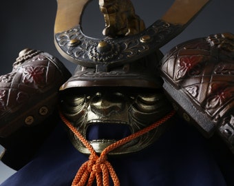 May include: A traditional Japanese samurai helmet, or kabuto, with a menacing face mask and intricate details. The helmet is made of metal and has a dark brown finish. The helmet is decorated with gold accents and a red and black floral pattern.