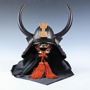 Könnte beinhalten: Schwarzer Samurai-Helm mit großen, gebogenen Hörnern und einem goldfarbenen Emblem. Der Helm hat einen goldfarbenen Gesichtsschutz und orangefarbene Quasten. Er liegt auf schwarzem Stoff. Traditionelle japanische Rüstung.