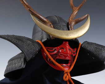 Puede incluir: Un casco de samurái negro con una máscara roja y cuernos. El casco tiene detalles intrincados y una luna creciente dorada en la parte delantera.