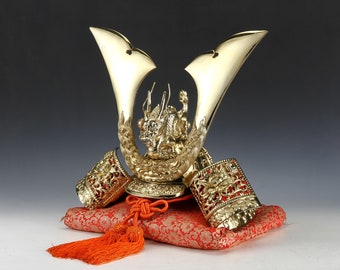 May include: A gold samurai helmet with a dragon design on top, resting on a red and gold cushion with a tassel.
