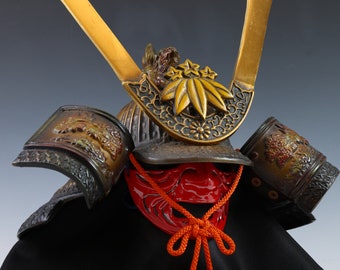 May include: A traditional Japanese samurai helmet, or kabuto, with intricate gold and black detailing. The helmet features a stylized leaf design and a red face mask. The helmet is displayed on a black stand.