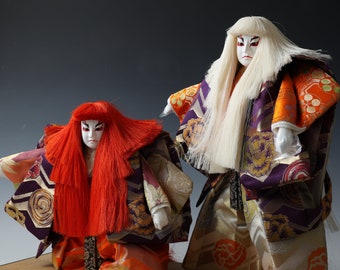 May include: Two traditional Japanese dolls with elaborate costumes. The doll on the left has red hair and a purple and gold patterned robe. The doll on the right has white hair and a purple, gold, and orange patterned robe.