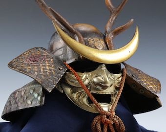 May include: A samurai helmet with a gold face mask, antlers, and a crescent moon brow guard. The helmet is decorated with intricate details and is made of metal.