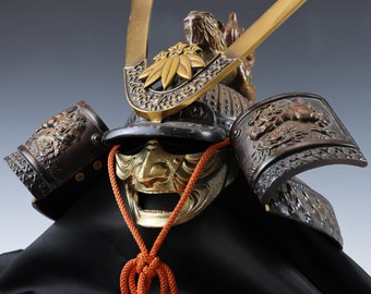 May include: A traditional Japanese samurai helmet, or kabuto, with a gold and black face mask, intricate details, and a red cord. The helmet is decorated with a dragon and floral motifs.