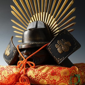 Puede incluir: Imagen detallada de un casco de samurái japonés, con acabado en bronce oscuro y detalles intrincados. El casco está adornado con una cresta dorada en forma de sol y descansa sobre un cojín con estampado rojo y dorado. Un cordón y borlas naranjas añaden color.