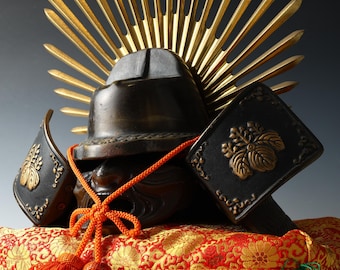 May include: A detailed image of a Japanese samurai helmet, featuring a dark bronze finish and intricate details. The helmet is adorned with a golden sunburst crest and rests on a red and gold patterned cushion. An orange cord and tassels add a pop of color.