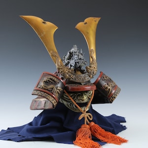 Puede incluir: Un elaborado casco de samur&aacute;i japon&eacute;s, con cuernos dorados y detalles intrincados de dragones. El casco presenta una combinaci&oacute;n de colores rojo y negro con detalles dorados y se exhibe sobre una tela azul, con borlas naranjas.