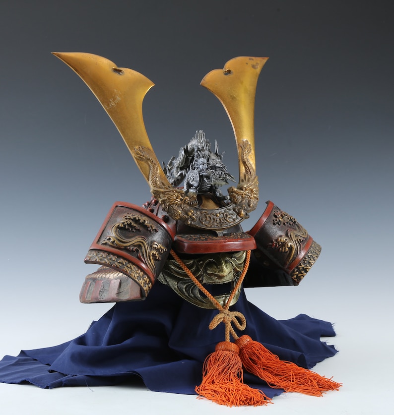 Puede incluir: Un elaborado casco de samur&aacute;i japon&eacute;s, con cuernos dorados y detalles intrincados de dragones. El casco presenta una combinaci&oacute;n de colores rojo y negro con detalles dorados y se exhibe sobre una tela azul, con borlas naranjas.
