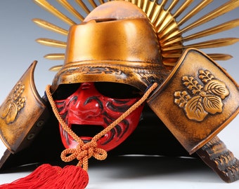 May include: A gold samurai helmet with a red face mask and a gold sunburst design. The helmet is decorated with a floral pattern and has a red tassel.
