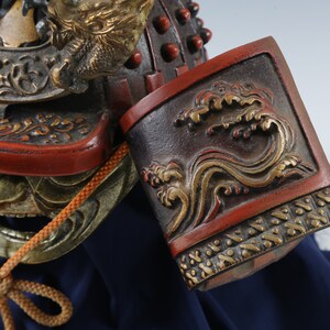Puede incluir: Primer plano de un casco de samur&aacute;i japon&eacute;s con detalles intrincados. El casco presenta una combinaci&oacute;n de colores rojo y negro con detalles dorados, incluyendo un motivo de drag&oacute;n y olas. Se ve un cord&oacute;n marr&oacute;n.