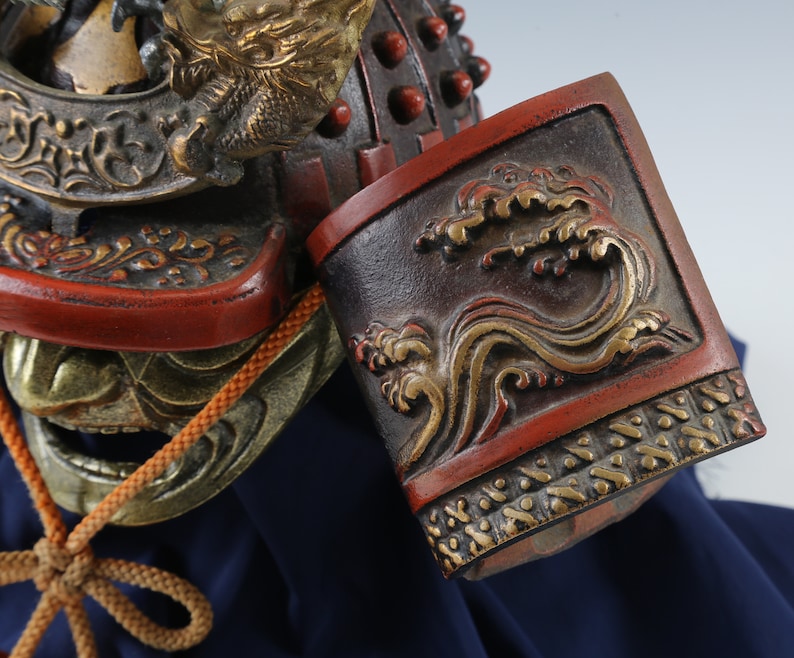 Puede incluir: Primer plano de un casco de samur&aacute;i japon&eacute;s con detalles intrincados. El casco presenta una combinaci&oacute;n de colores rojo y negro con detalles dorados, incluyendo un motivo de drag&oacute;n y olas. Se ve un cord&oacute;n marr&oacute;n.