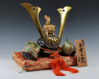 May include: A miniature samurai helmet with gold and black accents. The helmet is decorated with a gold lion and green and gold accents. The helmet is sitting on a red and gold cushion. The text on the wooden plaque reads "林 彦  光  忠  付  幅".