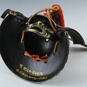 Puede incluir: Un casco de samur&aacute;i japon&eacute;s negro y dorado con una m&aacute;scara facial decorativa. El interior del casco es negro, con letras doradas. Cuerdas naranjas y una borla roja est&aacute;n unidas en la parte superior.