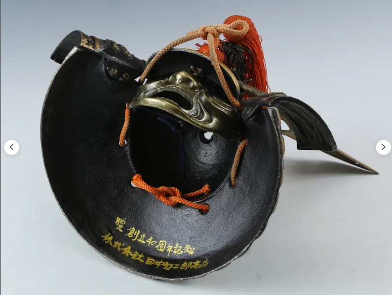 Puede incluir: Un casco de samur&aacute;i japon&eacute;s negro y dorado con una m&aacute;scara facial decorativa. El interior del casco es negro, con letras doradas. Cuerdas naranjas y una borla roja est&aacute;n unidas en la parte superior.