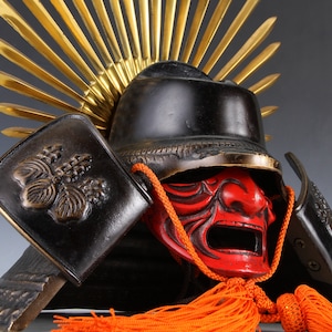 Puede incluir: Un casco de samurái japonés tradicional con una máscara facial roja, detalles dorados y un diseño negro y dorado. El casco está decorado con un patrón floral y tiene una larga borla naranja.