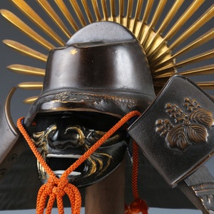 Puede incluir: Un casco de samurái negro con un diseño de sol radiante dorado en la parte superior. El casco tiene una máscara facial negra con detalles dorados y un cordón rojo.