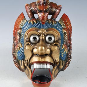 May include: A colorful wooden mask with a dragon on top and a small bowl hanging from its mouth. The mask is painted in shades of red, gold, blue, and green. The dragon is red and gold with a fierce expression.