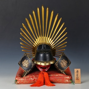 Puede incluir: Un casco de samurái negro y dorado con un diseño de sol naciente sobre un cojín rojo. El casco está decorado con detalles dorados y una máscara facial roja. El cojín tiene un patrón floral rojo y dorado. El texto en la placa de madera dice "秀光堂".