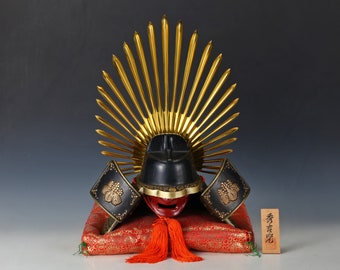 May include: A black and gold samurai helmet with a sunburst design on a red cushion. The helmet is decorated with gold accents and a red face mask. The cushion has a gold and red floral pattern. The text on the wooden plaque reads "秀光堂".