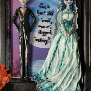 Peut inclure: Une œuvre d'art encadrée représentant un couple habillé en mariée et en marié. La mariée porte une robe blanche avec des accents bleus et une rose bleue. Le marié porte un costume noir avec une chemise blanche et une cravate noire. L'œuvre d'art comporte une citation : "Can a heart still break once it's stopped beating?"