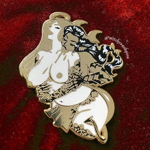 May include: Gold-tone enamel pin featuring a white angel and a black devil embracing. The angel has a halo and the devil has horns. The pin is on a red velvet background. @witchfsandpins