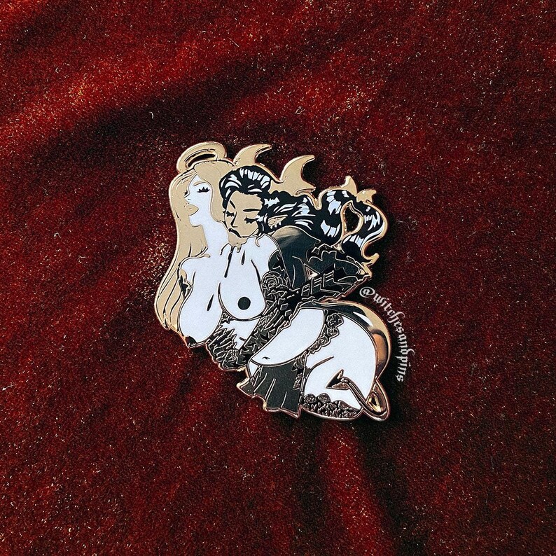May include: Gold-tone enamel pin featuring a black and white illustration of a woman with angel wings and a devil with horns. The woman is holding the devil's hand and they are both looking at each other. The pin has a black and white design with a gold outline. The text "@witchfressandpins" is on the bottom right corner of the pin.