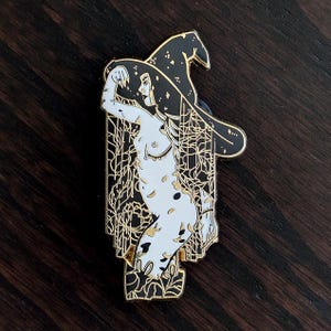 Könnte beinhalten: Ein schwarz-goldener Emaille-Pin mit einer stilisierten Illustration einer Hexe mit Hut. Die Hexe ist in Weiß mit goldenen Akzenten dargestellt, vor einem dekorativen Hintergrund. Der Pin hat ein detailliertes Design und ein glänzendes Finish.