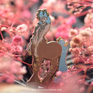 Puede incluir: Un pin de esmalte dorado que representa un centauro de pelo azul con una corona dorada. El centauro está de pie sobre sus patas traseras con sus patas delanteras levantadas. El pin está colocado sobre un fondo de flores rosas.