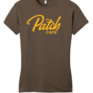 Pode incluir: Camiseta marrom com a inscrição "the Patch CAFE" em amarelo. O texto é escrito em itálico. A camiseta tem mangas curtas e gola redonda.