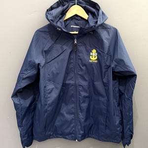 May include: Navy blue hooded jacket with a zipper closure. The jacket features a yellow emblem with the text "USN RETIRED" on the left chest. The jacket is hanging on a wooden hanger.