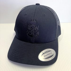 Könnte beinhalten: Marineblaue Trucker-Kappe mit Mesh-Rückseite und gebogenem Schirm. Auf der Vorderseite der Kappe befindet sich ein gesticktes Ankerdesign mit den Buchstaben "USN". Auf der Seite der Kappe befindet sich ein silbernes und schwarzes Etikett.