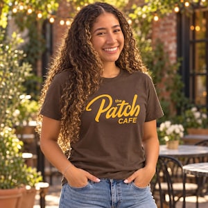 Könnte beinhalten: Ein dunkelbraunes Kurzarm-T-Shirt mit dem Schriftzug "the Patch CAFE", der in einer kräftigen gelben Schreibschrift und serifenlosen Schrift auf der Brust gedruckt ist. Das Shirt hat einen Rundhalsausschnitt und wird zu einer hellblauen Jeans getragen.