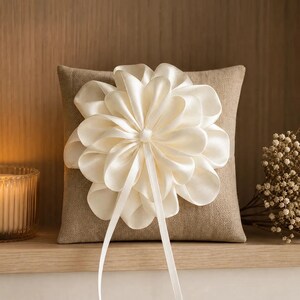 Peut inclure: Un coussin d'alliances carré en lin beige orné d'une grande fleur en ruban de satin crème à plusieurs épaisseurs au centre. Deux longs rubans de satin fins pendent du cœur de la fleur, ajoutant un détail décoratif sur le coussin.