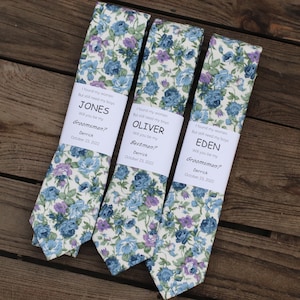 Peut inclure: Trois cravates avec un fond blanc et un motif floral dans des tons de bleu et de violet. Chaque cravate porte une étiquette blanche avec le texte "J'ai trouvé mes femmes mais j'ai toujours besoin de mes garçons" suivi d'un nom, "JONES", "OLIVER" ou "EDEN". Les étiquettes comprennent également le texte "Veux-tu être mon témoin ?" ou "Meilleur homme ?" ainsi que le nom "Derrick" et la date "23 octobre 2022".