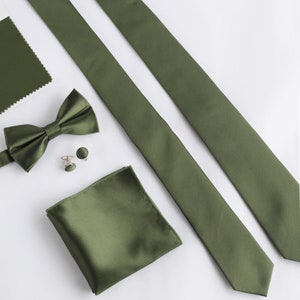 Könnte beinhalten: Olivgrünes Herren-Accessoire-Set, bestehend aus zwei Krawatten, einer Fliege, einem Einstecktuch und Manschettenknöpfen. Das Set ist auf einer weißen Oberfläche angeordnet, mit einem Stoffmuster in der oberen linken Ecke.