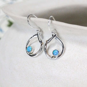 May include: A pair of silver dangle earrings featuring a blue opal gemstone in each earring. The earrings are shaped like a teardrop with a circular opening.