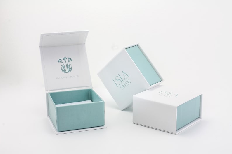 Puede incluir: Cajas de joyer&iacute;a blancas con interior azul claro y cierre magn&eacute;tico. Una caja est&aacute; abierta, revelando el interior. Las cajas tienen el texto "ISLA SILVER" y un dise&ntilde;o floral.