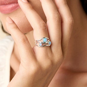 May include: A silver ring with three opal stones. The ring has a unique, flowing design. The opals are light blue and round. The ring is on a person's finger. A matching earring is visible in the background.