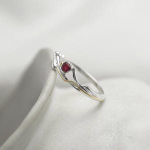 May include: A silver ring with a delicate, twisted design featuring a small, red gemstone. The ring is set against a white background.