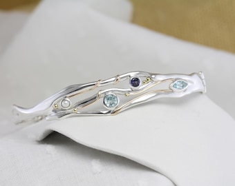 May include: A silver-tone bracelet with gold accents and inlaid gemstones. The bracelet features a flowing, organic design with a pearl, a dark purple stone, and two light blue stones. The bracelet is resting on a white surface.