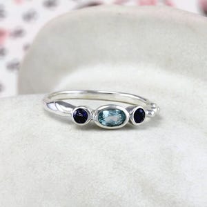 May include: A silver ring with three gemstones. The center stone is a light blue oval, and the two side stones are dark blue rounds.