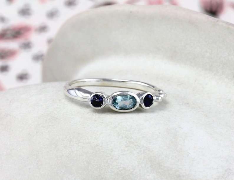 May include: A silver ring with three gemstones. The center stone is a light blue oval, and the two side stones are dark blue rounds.