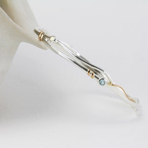 以下が含まれることがあります： 金色のアクセントと、小さく丸い水色とオフホワイトの宝石が付いた、銀色のバングルブレスレット。 ブレスレットは波状のデザインで、白い背景に配置されています。