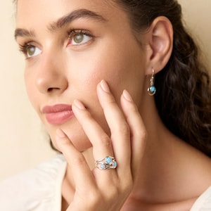 May include: Silver ring with three light blue opal stones. The ring is on a woman's finger. Matching earrings with a single light blue stone are also visible. The jewelry is set against a soft, neutral background.