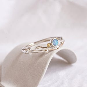 May include: A silver ring with a light blue opal stone and a small gold accent. The ring has a unique, organic design with a textured band. The opal is round and set in a delicate silver setting. The ring is displayed on a neutral surface.