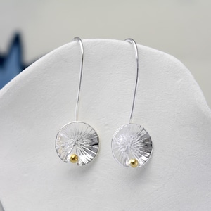 May include: Silver drop earrings with a textured, circular design. Each earring has a small, gold-coloured bead at the centre. The earrings are displayed on a white surface, with a blurred blue and white background.