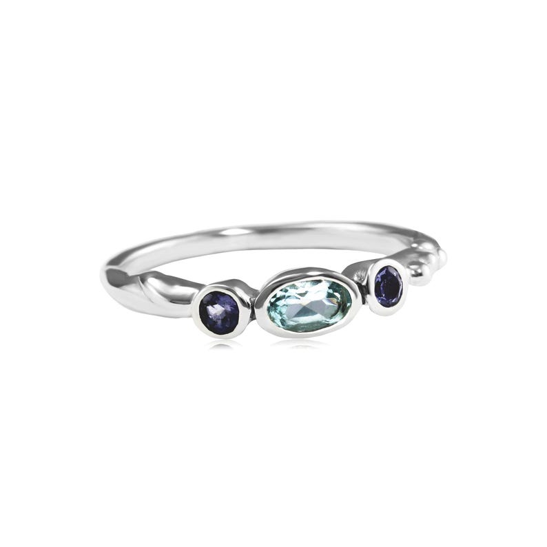 May include: A silver ring with three gemstones. The center stone is a light blue oval, and the two smaller stones are dark purple and round.